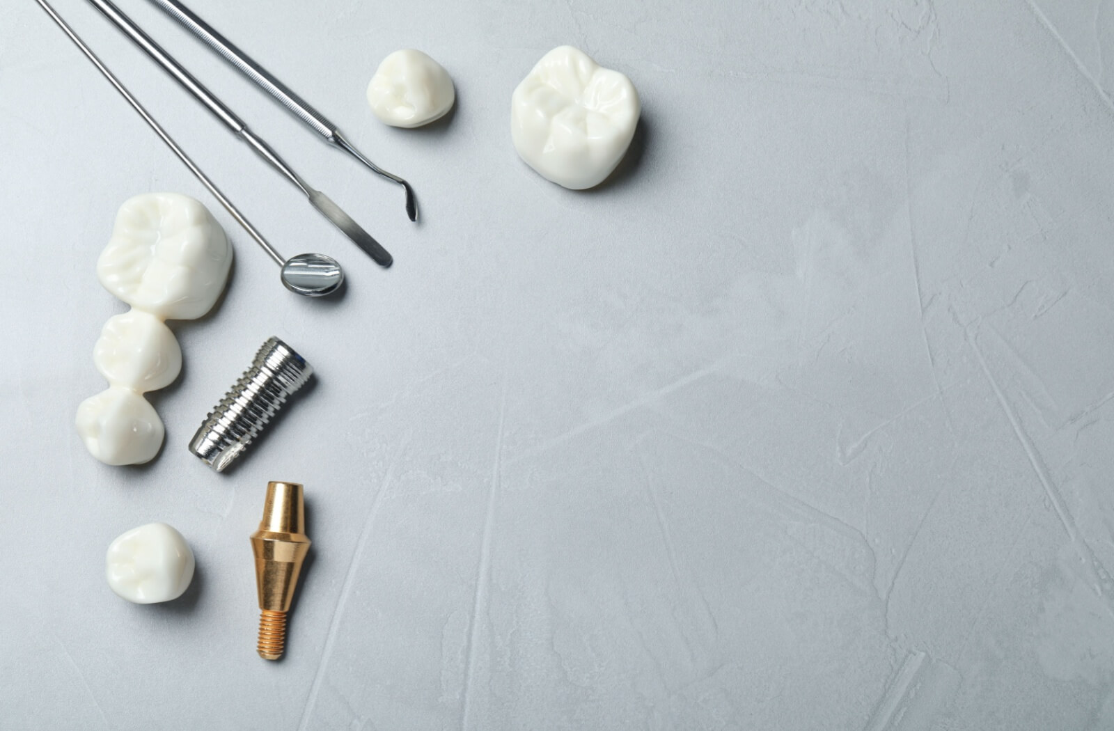 Dental tools sit beside a dental implant screw, the abutment that goes with it, and an assortment of dental crowns in many styles like molars, front teeth, and multiple teeth on one bridge.