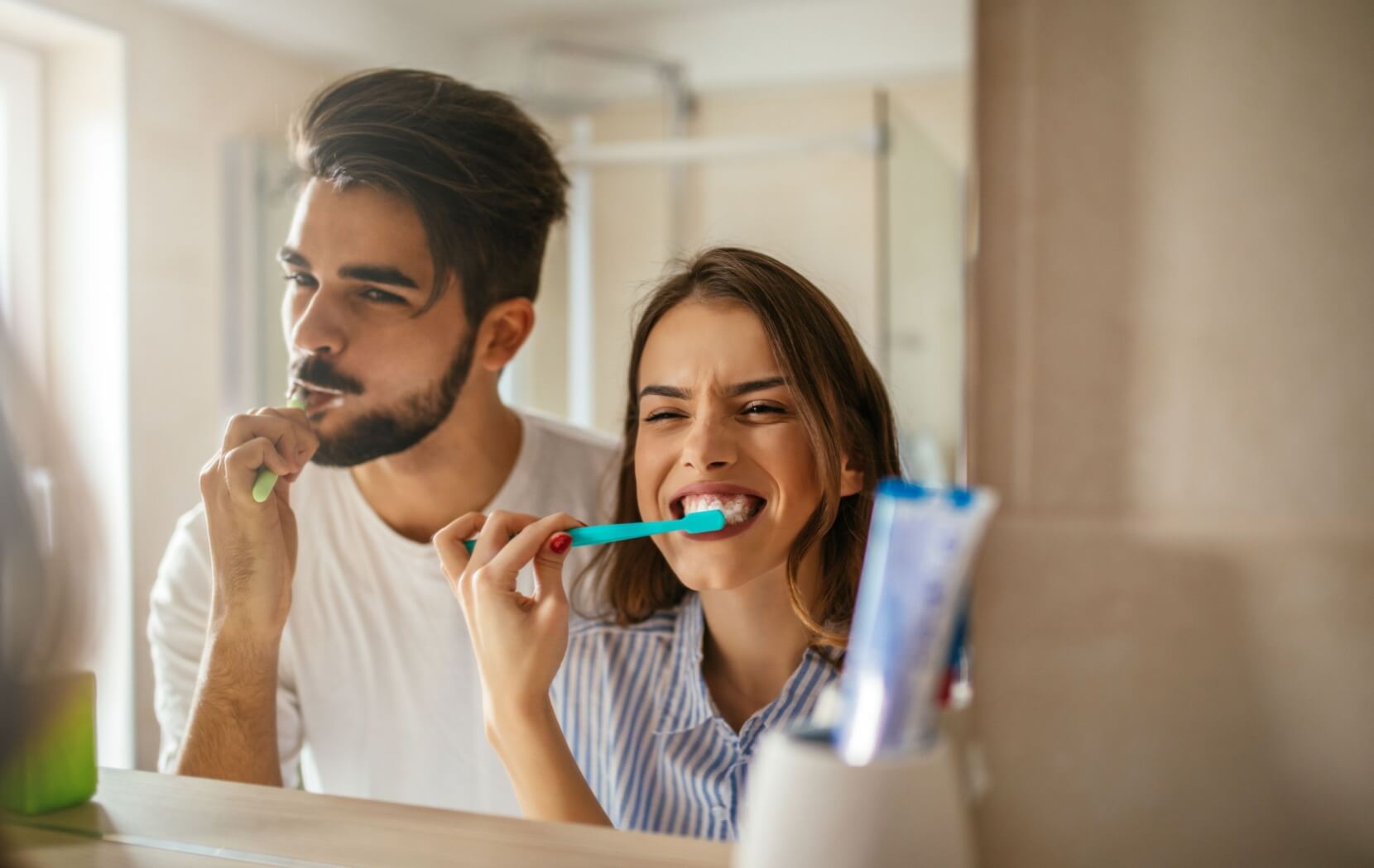A happy young couple brush their teeth together in the morning in front of their bathroom mirror.