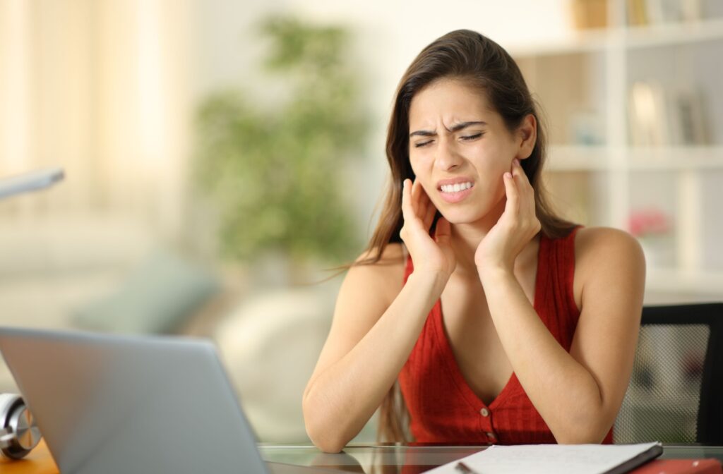 Woman sitting at a desk in front of a computer, rubbing her jaw, experiencing a TMJ headache.