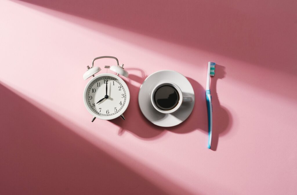 Alarm clock, cup of coffee, and toothbrush on a plain pink background.