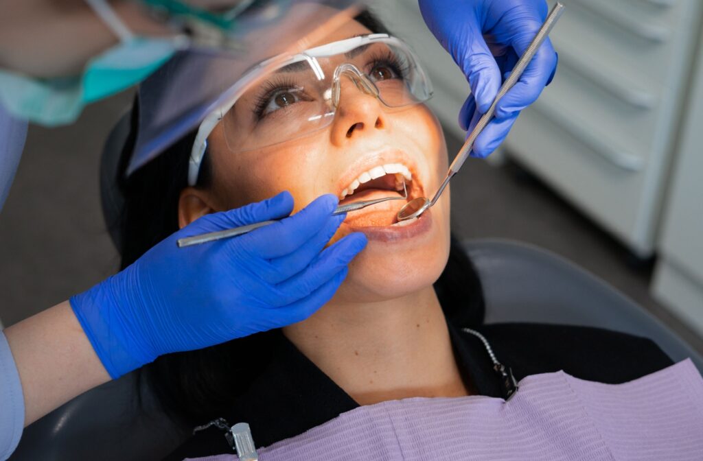A person laying in a dental chair, mouth open, having a dental hygienist clean their teeth during a routine dental visit.