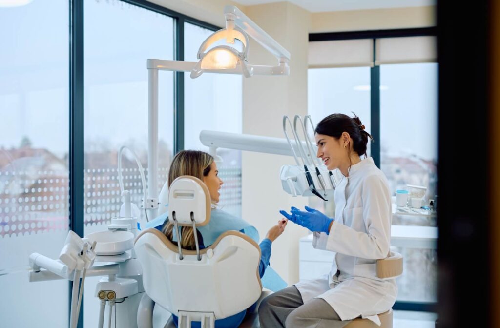 A dentist in a white lab coat and blue gloves talking to a patient in a dental chair in a brightly lit modern office.