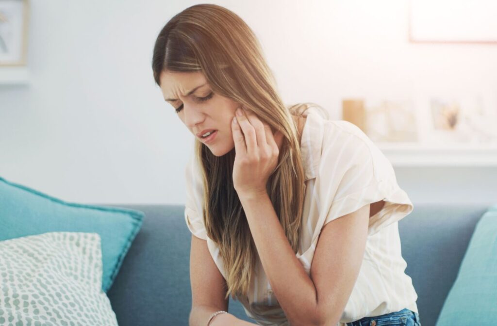 A person sitting on a couch wincing and holding her cheek in pain, illustrating the discomfort often associated with wisdom teeth.